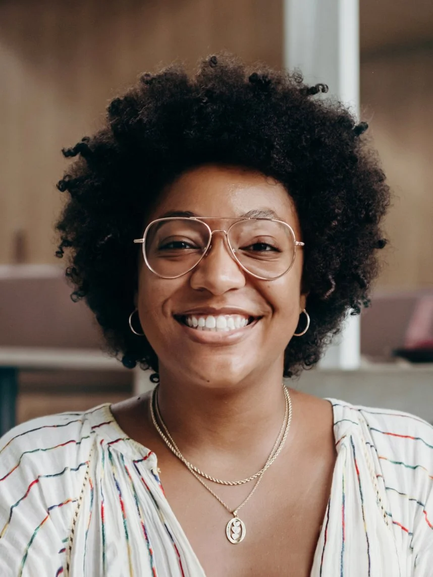 A woman smiling with glasses, star-shaped earrings, layered necklaces, and a striped blouse in an indoor setting.
