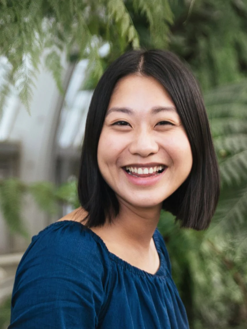 Smiling woman with shoulder-length black hair wearing a blue top outdoors with green foliage in the background.