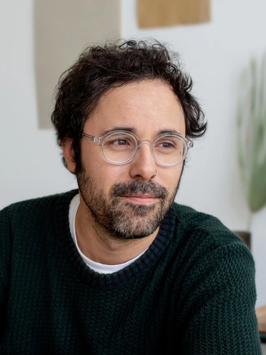 A man with curly dark hair, glasses, and a beard wearing a dark green sweater, sitting indoors in front of a blurred background.