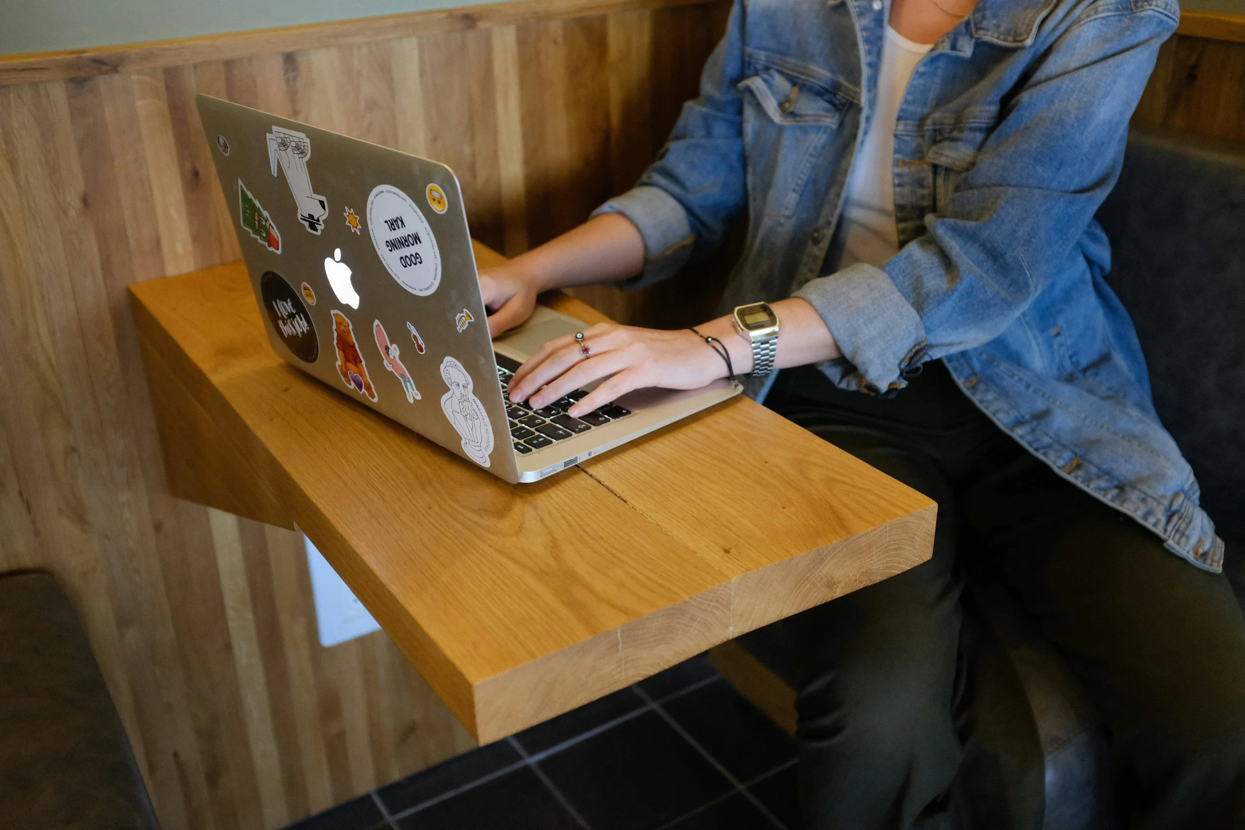 Person in a denim jacket using a laptop with various stickers, sitting at a wooden table in an office.