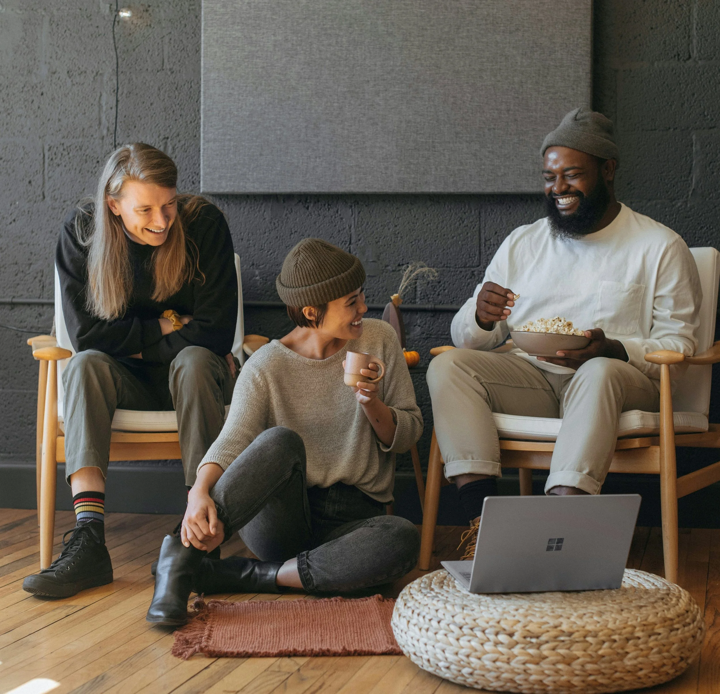 Three coworkers enjoying a video conference in an office, one sitting on a floor mat with a laptop, another sitting on a chair with a bowl, and the third sitting on a chair, all smiling and sharing a happy moment.