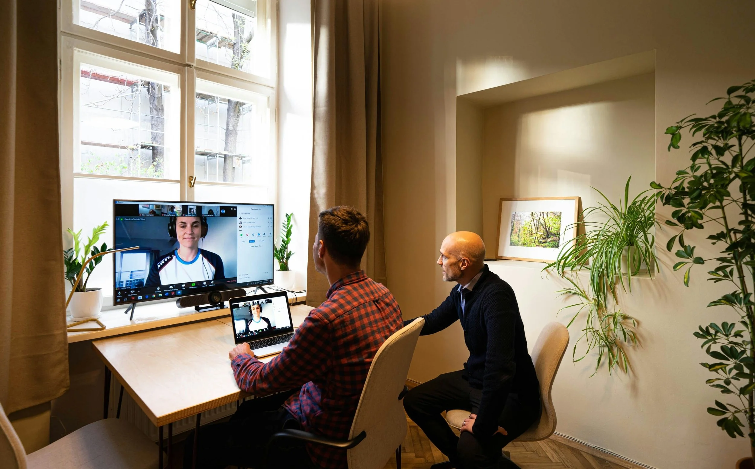 Two men in a room sitting at a desk, participating in a video conference call on a large monitor and laptop, with a woman smiling on the screen, plants by the window, and framed pictures on the wall.
