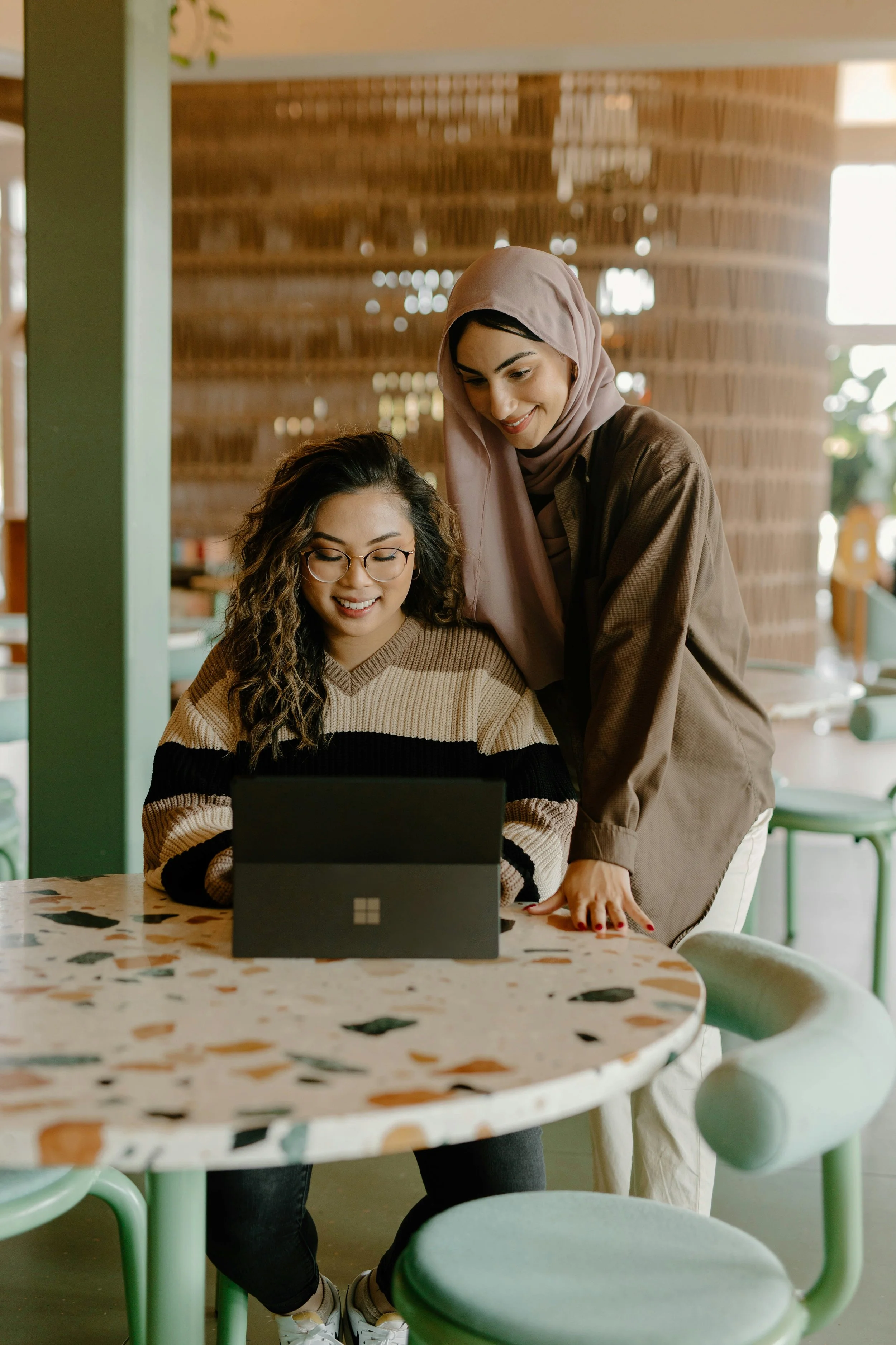 Two women look at a laptop together in a modern cafe with a terrazzo table and green chairs, one wearing glasses and the other wearing a hijab.