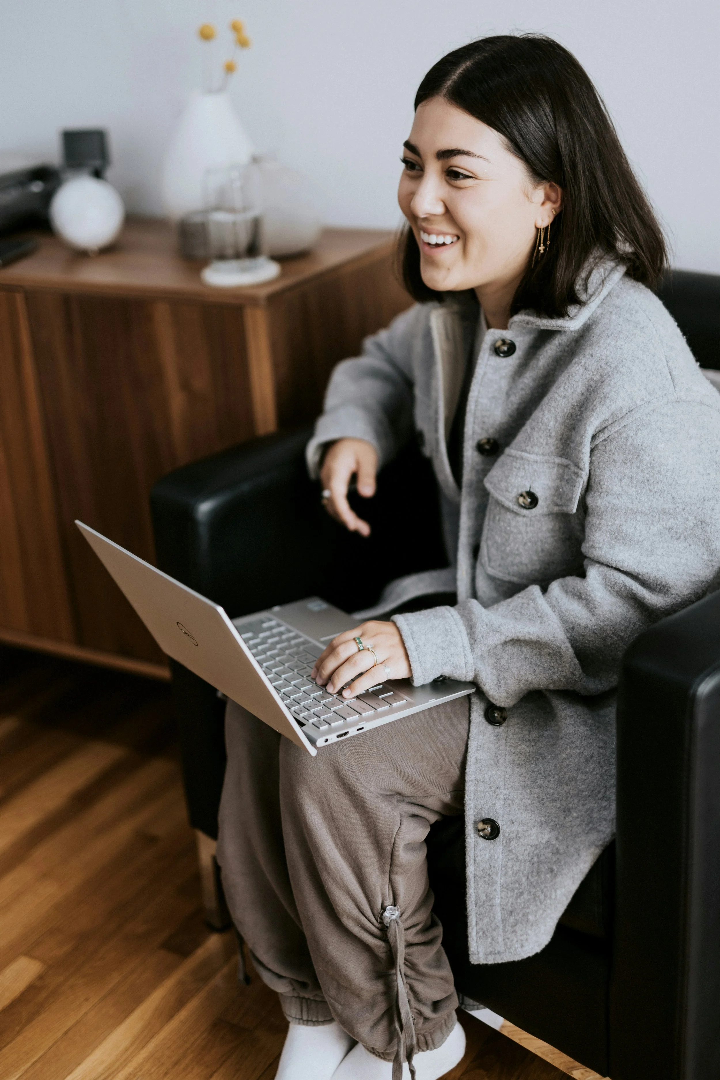 A woman with dark hair and earrings smiling while sitting in a black chair, using a laptop on her lap in an office with wooden furniture and a white wall.